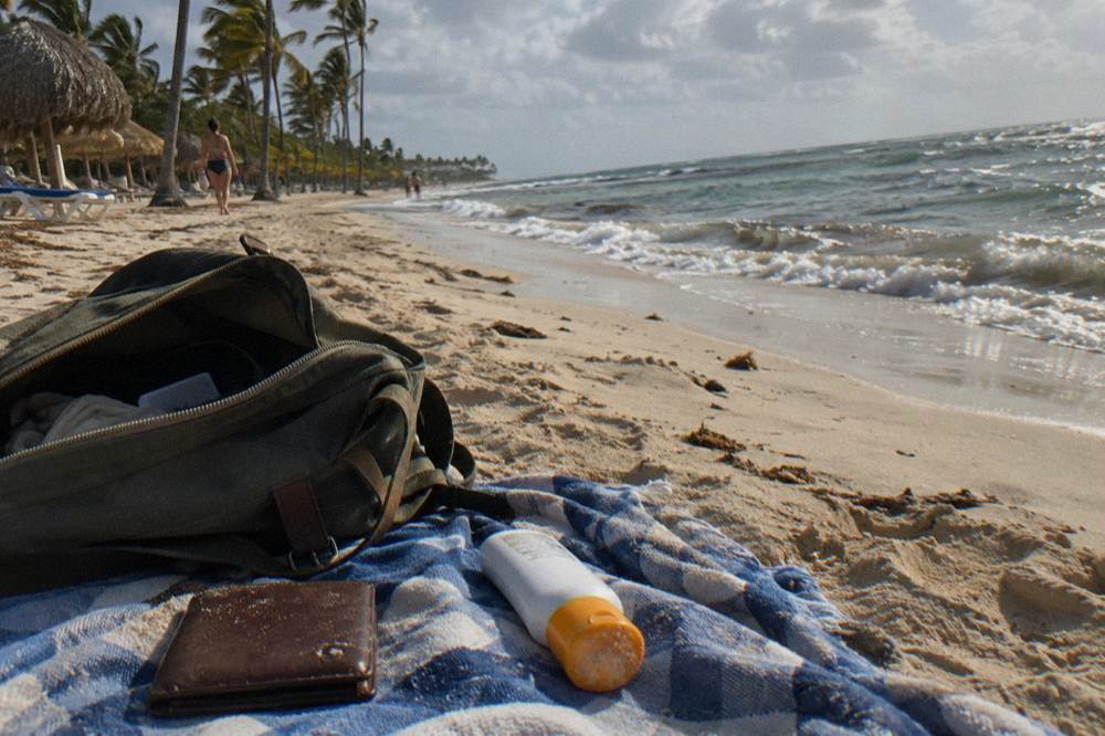 A travel wallet and sunscreen resting on a beach towel beside an open daypack
