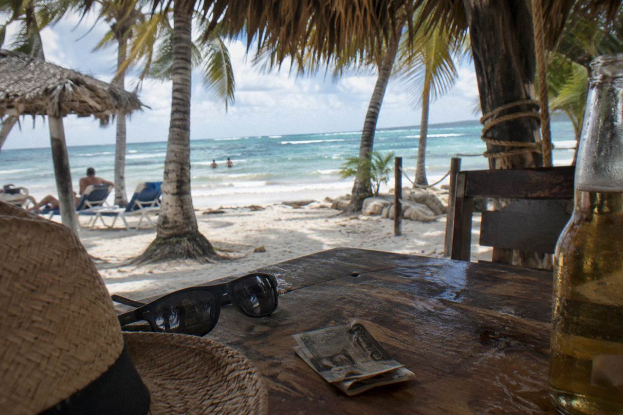 Shaded beach bar table with sunglasses, a straw hat and small dollar bills set beside a cold drink