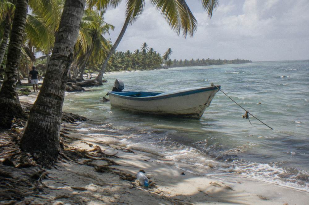 A shaded palm-lined beach with calm water and a moored small boat