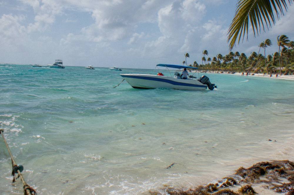 Turquoise shallow water over a sandbar with a speedboat anchored nearby and scattered palm trees on the horizon