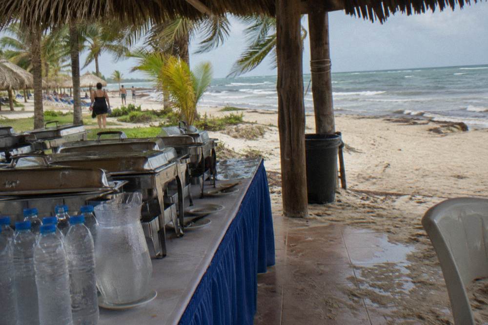 A beach buffet table with covered dishes and bottled drinks under a palm-thatch shelter