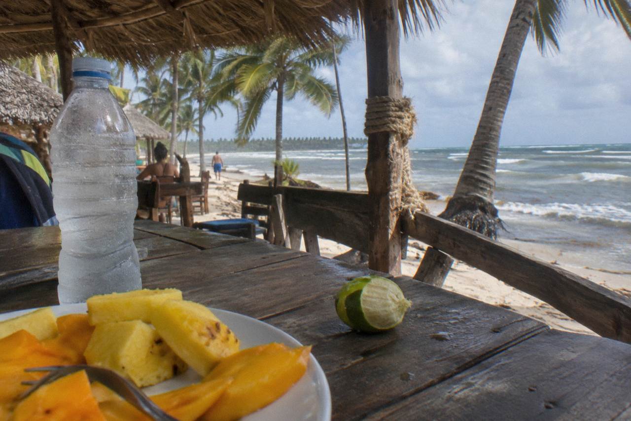 Fresh tropical fruit and bottled water arranged on a wooden table beside an open-air restaurant