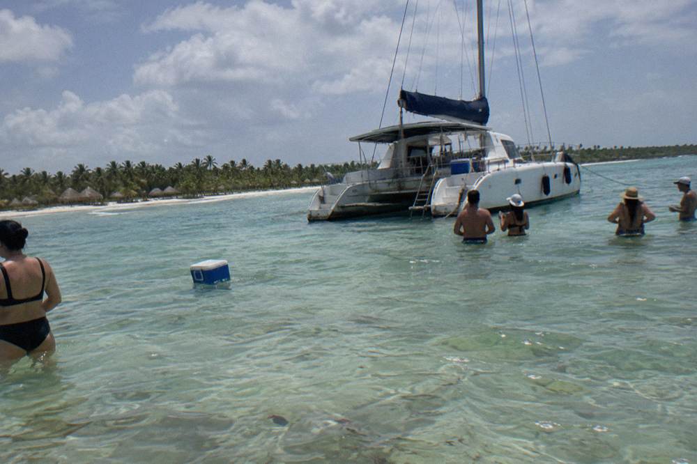 Catamaran anchored in shallow clear water beside a sandbar with guests standing in the sea