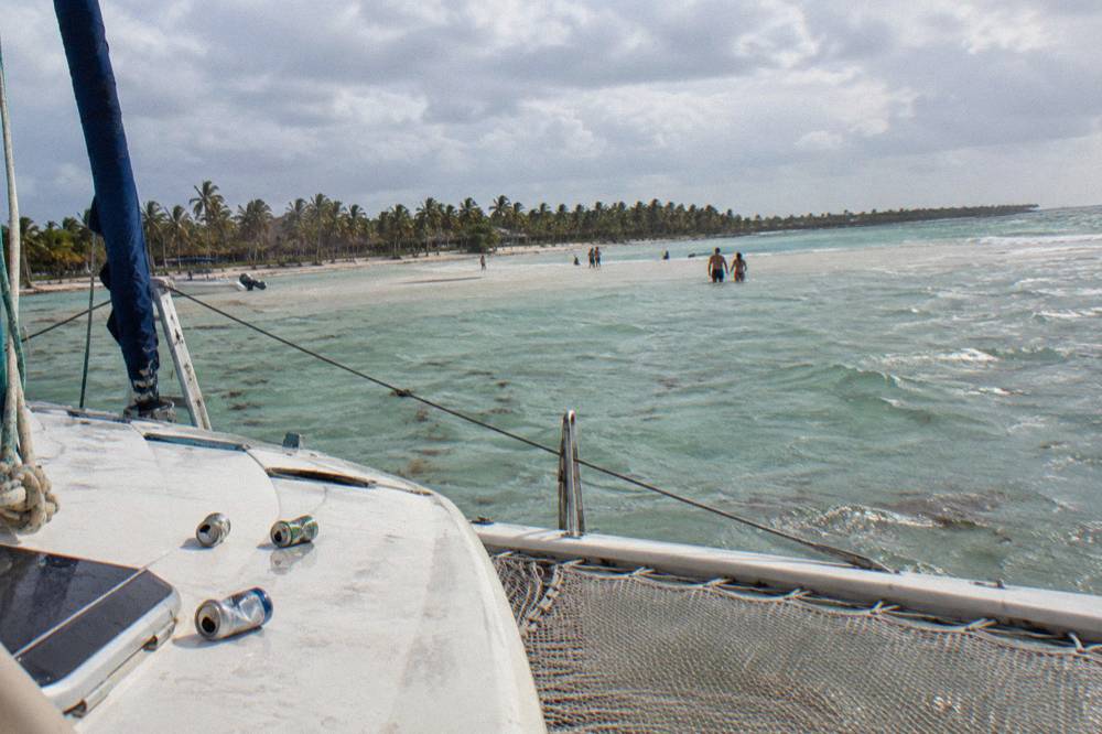 A catamaran anchored near a shallow sandbar with clear water and palm trees on the horizon