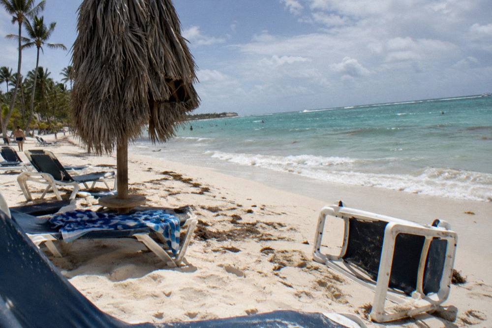 Beach chairs and a palm thatched umbrella on white sand under a clear sky