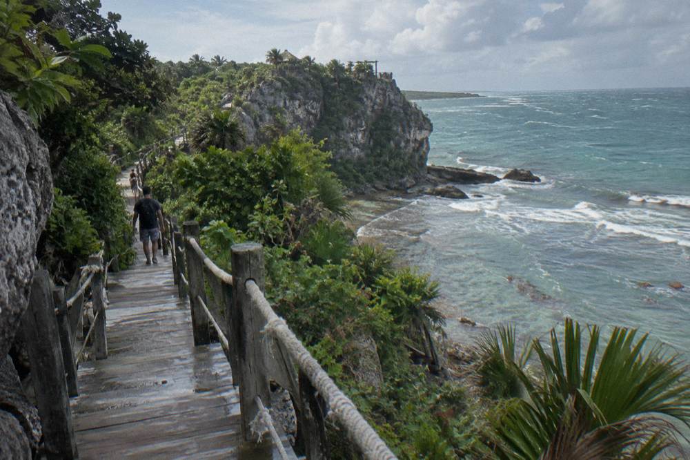 Wooden walkway through tropical forest leading toward a limestone cliff and blue water