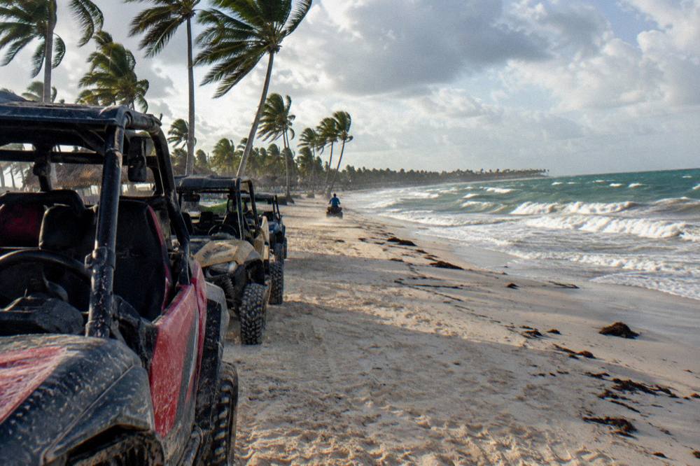 A line of buggies parked on pale sand at a wide open-ocean beach with rolling waves