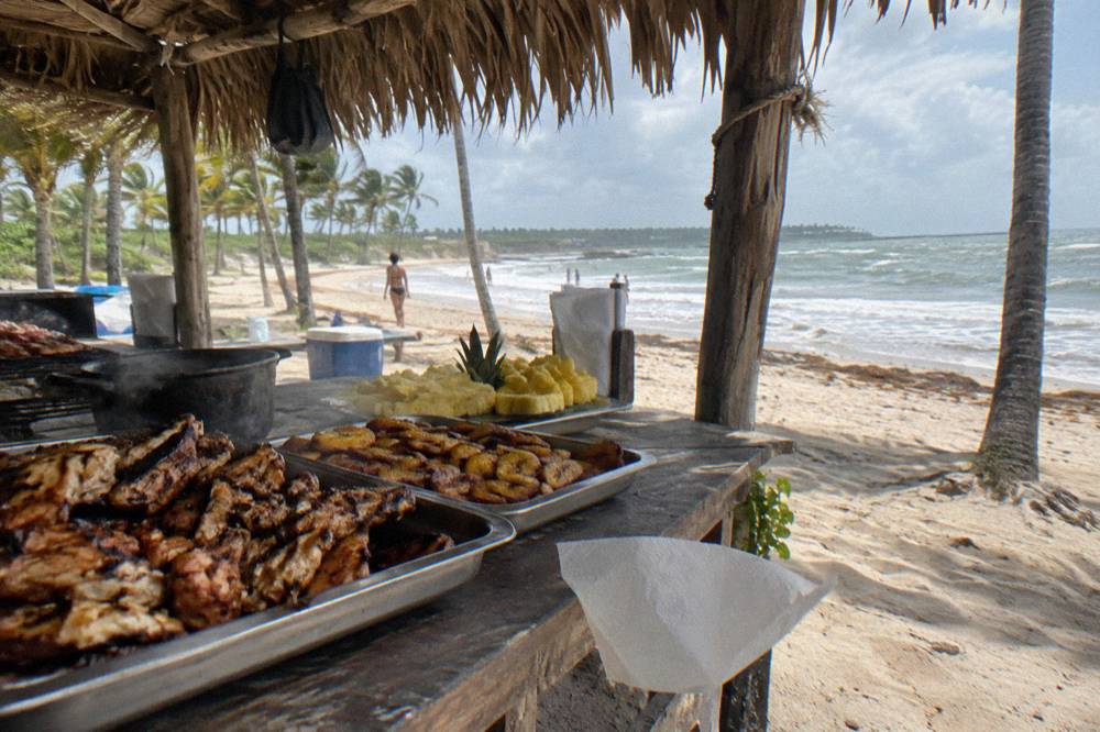 A beachside buffet table with grilled food and tropical fruit under a palm thatch roof