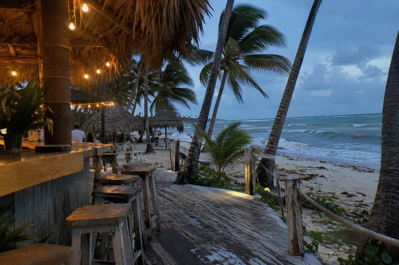 Open-air beach bar with string lights glowing above palm trees and empty stools along a wooden deck at dusk