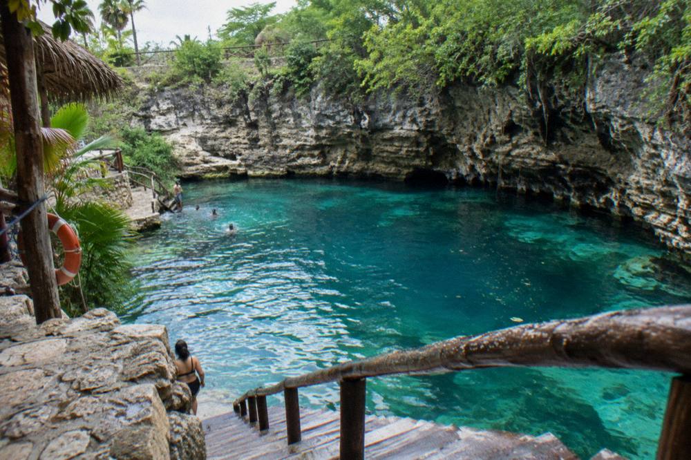 A turquoise cenote surrounded by rock walls and wooden steps leading to the water