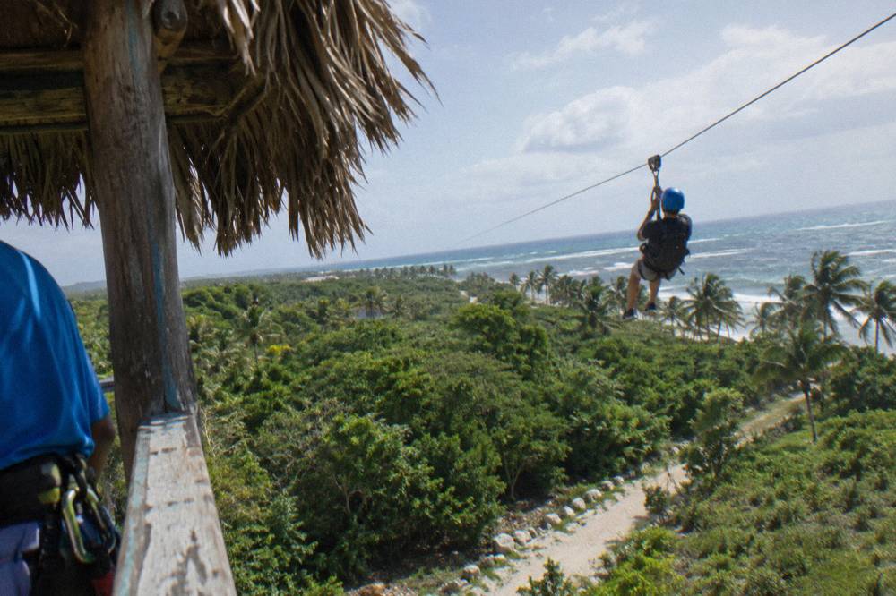 Rider in a helmet and harness gliding along a cable above tropical treetops