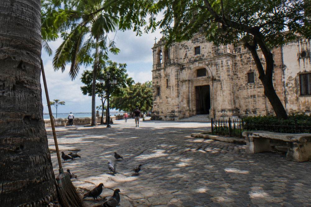 A sunlit colonial plaza with an old cathedral facade and shade trees