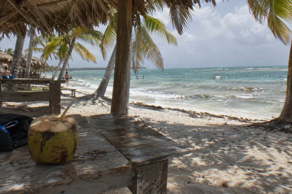A shaded beach lounge area with wooden benches and palm thatch roofing on white sand